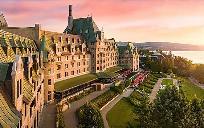 Hotel de estilo castillo con techos de cobre verde, buhardillas y terrazas escalonadas sobre un jardín, al atardecer, con el río al fondo.